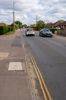 Norwich, UK- 17 Mayıs 2025: Huzurlu banliyö caddesi, hareket halindeki araçlar ve tuğlalı evler görünür. Olgun ağaçlar, çalılar ve sarı merkez çizgileri olan sessiz bir mahalle yolu..