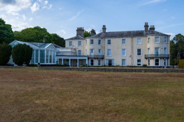 Norwich, UK- 17 Mayıs 2025: Dönem mimarisi ve eklenmiş glasshouse konservatuvarı olan büyük tarihi malikane. Manikürlü çimenler ve etrafını çevreleyen olgun ağaçlar içinde zarif, krem rengi bir ev.