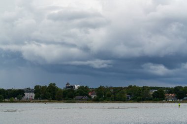 Tallinn, Estonya - 21 Ağustos 2025: yoğun yeşil yeşillik ve kıyı şeridi ile deniz kenti üzerinde fırtına bulutları toplanıyor. Yerleşim alanının üzerindeki gri gökyüzü ve sakin sularla atmosferik kıyı manzarası