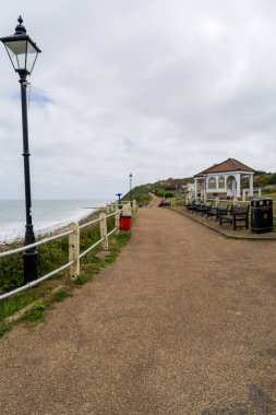 Cromer, UK- 18 Mayıs 2025: Eski bir sokak lambası ve deniz kenarındaki uçurum boyunca uzanan banklar ve korkuluklar ile okyanus kıyı geçidine bakan beyaz çardak köşklü deniz kenarı geçidi.