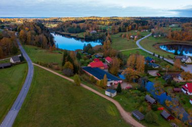 Rouge, Estonya - 17 Ekim 2020: Sonbaharda yansıtıcı bir göl yakınlarındaki sakin bir kırsal yerleşim yerine kuş bakışı bakış. Dönek yollar dağınık evleri renkli yaprak döken ağaçlar arasında birleştiriyor.