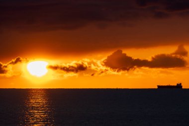 Ufukta büyük bir kargo gemisinin silueti, dramatik bir turuncu ve altın gökyüzü ve kara bulutlar altında, Baltık Denizi 'nde sersemletici bir gün batımı.