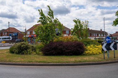 Norwich, UK- 17 Mayıs 2025: Dramatik, bulutlu bir gökyüzü altında tuğladan evler ve dükkanların yer aldığı yerleşim alanındaki renkli çalılar ve ağaçlarla döşenmiş trafik kavşağı