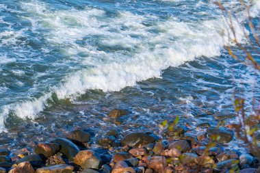 Köpüklü beyaz dalgalar kayalık kıyı şeridinde kayalık taşların ve çakıl taşlarının üzerine çarpıyor. Sahilde doğal su hareketleri ve doku kalıpları ile dinamik sıçrama ve sörf hareketleri yaratıyor.