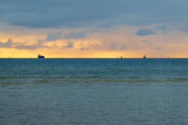 Ufukta kargo gemileriyle gün batımı deniz manzarası, sakin okyanus suyunun üzerindeki bulutlarla altın akşam gökyüzü, uzaktaki deniz gemilerinin siluetleri huzurlu bir kıyı manzarası yaratıyor.