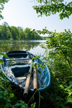 Eski mavi ahşap bir tekne, yazın yeşil ağaçlar ve yapraklarla çevrili, huzurlu göl kıyısına demirlemiş. Kırsal kesimde sakin suda geleneksel kayıkla sakin doğa sahnesi