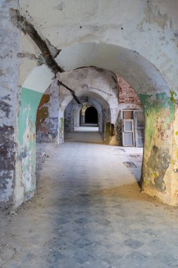 Terk edilmiş hapishane koridoru, kemerli tavan ve çürüyen duvarlara yeşil boya dökülüyor. Hapishane koridorunun iç kısmı birden fazla kemer yolu, hasarlı alçı ve kentsel bozulma gösteriyor.