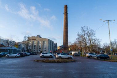 Tallinn, Estonya - 20 Aralık 2025: Yenilenmiş endüstriyel depo binası, kemerli pencereler ve güneşli mavi gökyüzünün altında modern park alanı olan eski fabrika bacası