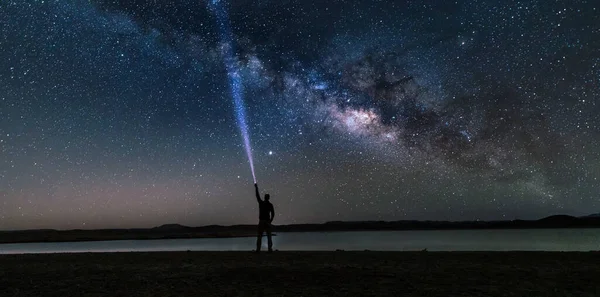 Astrophotography: Night sky starring the milky way over a man with a torch light standing at the shore of a lake with calm water surface, Morocco