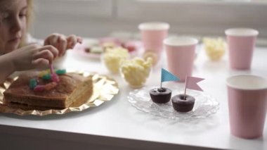 Side view of cheerful little kid in casual clothes sitting at wooden table near window and eating sweet cake during holiday