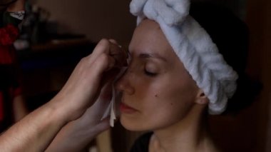 Kırpılmış suratsız makyaj sanatçısı, pamuklu kumaş çubuğunda göz makyajını temizliyor. Sakin, gözleri kapalı ve beyaz saç bandı takmış genç bir bayan müşteri.