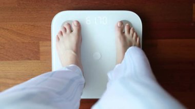 Üstten görünüşlü, çıplak ayaklı, rahat pijamalı, dijital ağırlığı ve vücut yağı terazisi üzerinde duran ve sabah banyoda 60 kg ağırlığında sağlıklı ağırlığı gösteren çıplak ayaklı bir kadın.