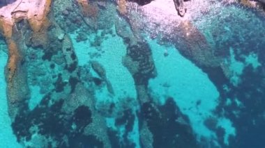 Güneşli bir günde, dalgalı kayalık kıyılarda görünen deniz yatağı olan turkuaz berrak deniz suyunun insansız hava aracı görüntüsünden.