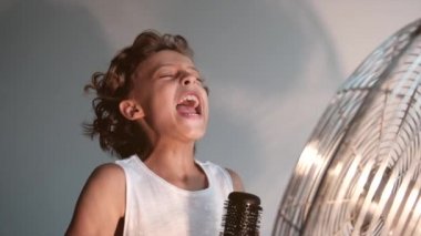 Duygusal yüz ifadesi olan bir çocuk, odanın içindeki kılları çıkaran bir pervaneyle masanın önünde bir mikrofon gibi şarkı söylüyor.
