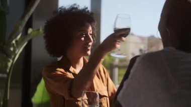 Kafede oturup kırmızı şarap içen ve kısa saçlı, tanınmayan bir arkadaşla konuşan neşeli, kıvırcık saçlı Afro-Amerikan kadın.