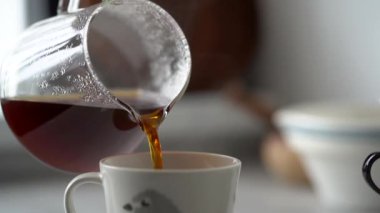 Anonim bir kadın sıcak demlenmiş kahveyi evdeki rahat mutfakta cam sürahide bardağa dolduruyor.