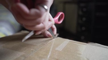 Evde paketleri açarken, mühürlü karton kutunun üzerindeki yapışkan bandı kesmek için makas kullanan isimsiz bir kişinin görüntüsü
