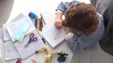 Maskeli çocuk karantina sırasında masada oturup internetten ders çalışırken not defterine not alıyor.