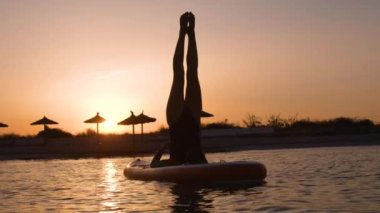 Gün batımında sörf tahtasında yoga yapan kadın sörfçü.