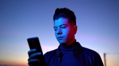 Siyah ceketli, ciddi bir modern adam, arka planda koyu mavi gökyüzü ile kırmızı ve mavi ışıkta dikilirken akıllı telefondan konuşuyor.