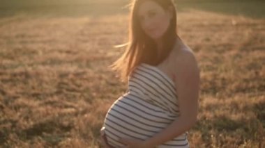 Tüm vücudu çizgili elbiseli mutlu hamile bir kadın karnına dokunuyor ve kırsaldaki çayırlarda kuru çimlerde dururken kameraya bakıyor.