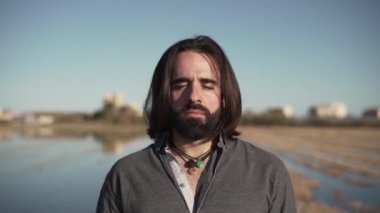 Göl kenarında gözleri kapalı duran uzun saçlı adam.