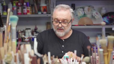 Ressam, dağınık bir masada oturup yaratıcı atölyede çalışırken el fırçasıyla yelpaze çiziyor.