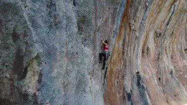 Güneşli bir günde yüksek dağın kayalık yamacında tırmanan halat ve güvenlik ekipmanlarıyla aktif kadın dağcı drone görüntüsünden.