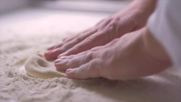 ピッツェリアキッチンで作業しながら 小麦粉とテーブルの上にピザのための生地を形成する作物シェフの遅い動きを閉じます — ストック動画