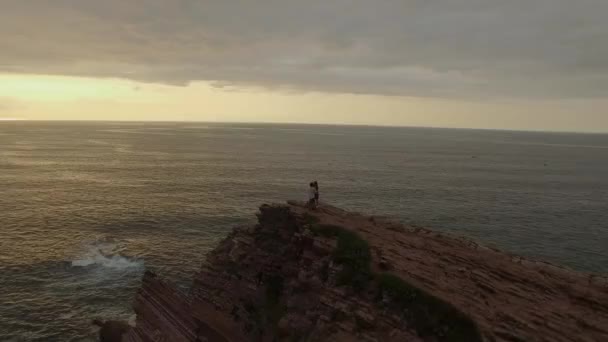 夕日で海に対して荒い岩の崖の端に立っている間に認識できないロマンチックなカップルキスの上からドローンビュー — ストック動画