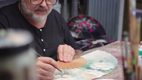 画家坐在凌乱的桌子前 在创意作坊工作时 用画笔在手扇上画画 — 图库视频影像