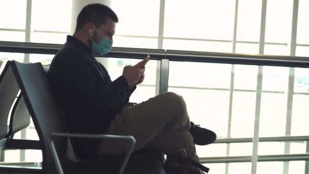 机场候机室内穿着休闲装 戴口罩 头戴智能手机 背靠背坐在椅子上的男性旅客在验尸官流行病流行期间的侧视图 — 图库视频影像