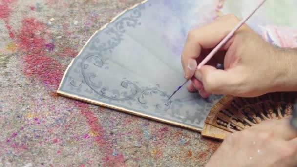 画家坐在凌乱的桌子前 在创意作坊工作时 用画笔在手扇上画画 — 图库视频影像