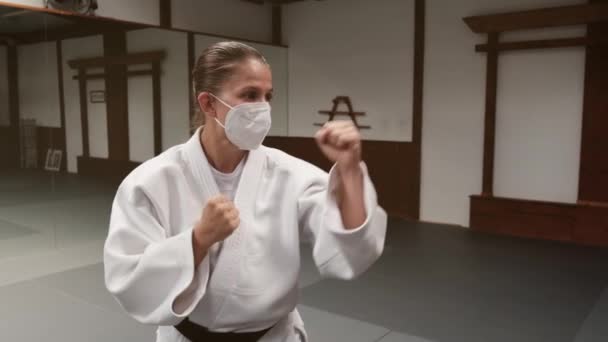 白い着物を着て 黒いベルトで保護顔のマスクを身に着けている編組の女性は 武道ジムで拳を握り締めたパンチで縛られています — ストック動画