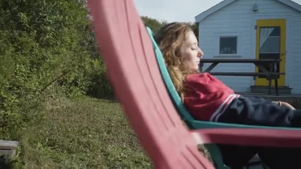 ニュースコットランド カナダの風の強い日に田舎のキャビンの庭で休んでいる間 ラウンジャーで寝ているカジュアルな服の若い女性のパンの右側のビュー — ストック動画