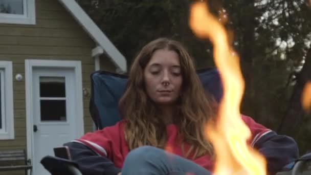 若い女性が椅子に座って カナダのニュースコットランドの夜にコテージの庭でたき火の近くでリラックス — ストック動画
