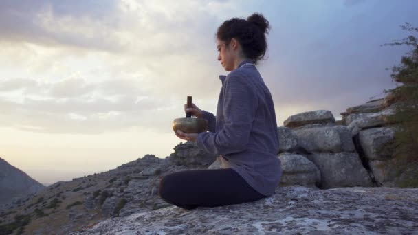 歌のボウルと瞑想しながら 日没時に山の中でリラックスしてパドマサーナの位置に座って静かな女性の低角度側のビュー — ストック動画