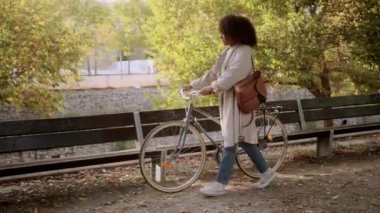 Kıvırcık saçlı, şık Afrikalı Amerikalı bir kadının park yolunda eski moda bir bisikletin yanında durup sonbahar gününün tadını çıkarırken görüntüsü.