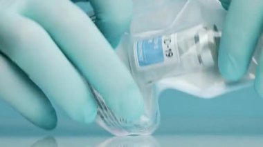 Lateks eldivenli isimsiz bir doktor COVID 19 'un mavi arka planındaki aşısıyla dolu cam şişeyle plastik torba açıyor.