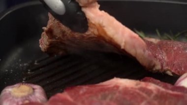 Mutfak maşası olan isimsiz bir aşçı ızgara tavada sulu et bifteğini biberiye ve ağır çekimde sarımsak ile çeviriyor.