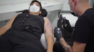 Kuafördeki masada yatan kadının kolunda makine dövmesi yapan bir erkek dövmeci.