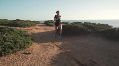 Siyah aktif giysili sporcu kadın huzurlu günbatımında yeşil deniz kıyısında koşuyor.