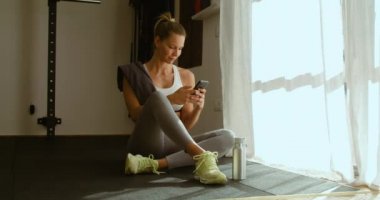 Spor giyim bölümünde yüksek açılı sakin bir kadın bir şişe suyla yerde oturuyor ve eğitimden sonra akıllı telefonlara bakıyor.