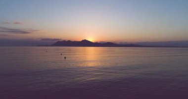 İnsansız hava aracı günbatımında sakin deniz yüzeyinde küçük adanın siluetine doğru uçarken karanlık sulara yansıyan güneş ışığı...