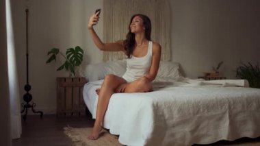 Young smiling female in shirt and panties using modern smartphone while sitting on cozy bed in light bedroom