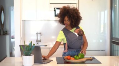 Afro-Amerikalı anne, mutfakta taze sebze kesip askıda uyuyan bebeği taşırken laptopta lezzetli bir yemek tarifi hazırlıyor.