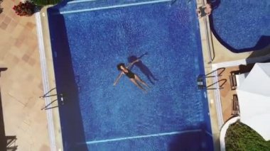 Yüzme havuzunda şeffaf suyun üzerinde uzanmış mayo giyen sakin kadın turistin yaz tatilini tatil köyünde geçirdiği drone görüntüsünü büyüt.