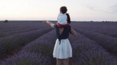 Omuzlarında küçük bir çocuk olan, mor lavanta çiçekleriyle çiçek açan ve günbatımının tadını çıkaran, tanınamayan, sevgi dolu bir annenin arka görünüşü.