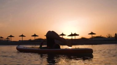 Gün batımında sörf tahtasında yoga yapan kadın sörfçü.