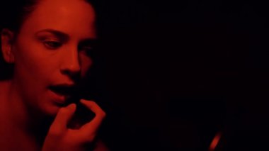 Siyah arka planda kırmızı ışıkla karanlık stüdyoda ruj sürerken konsantre olmuş çekici bir kadının yan görüntüsü.